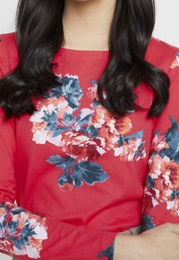 Punainen pitkähihaine paita, jossa on vaaleanpunaisia, valkoisia ja sinisiä kukkakuosia. Materiaali näyttää pehmeältä ja ribatulta.