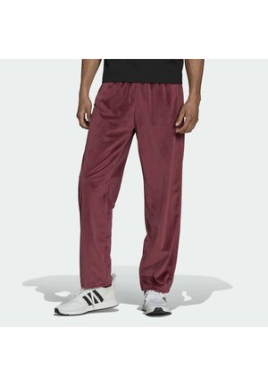 Homme portant un pantalon ample bordeaux et des baskets blanches à rayures noires, debout devant un fond clair uni.