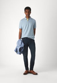 Polo blu chiaro con colletto giallo a contrasto, jeans scuri e mocassini marroni. Tiene una giacca blu su un braccio.