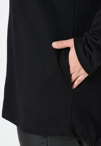 Tunic nera realizzata in morbido tessuto, con una tasca laterale e orlo arrotondato. Le maniche lunghe hanno polsini aderenti, offrendo una vestibilità rilassata.