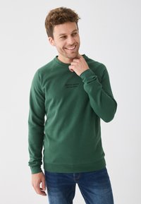 Sweatshirt vert avec manches longues, encolure ronde et poignets côtelés. Présente un texte sur le devant et un tissu de épaisseur moyenne. Porté avec un jean bleu.