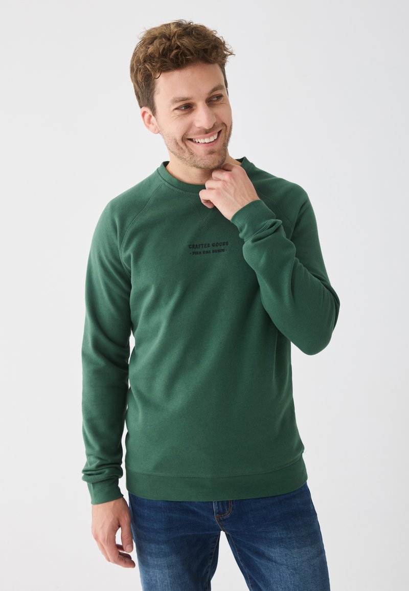 Sweatshirt vert avec manches longues, encolure ronde et poignets côtelés. Présente un texte sur le devant et un tissu de épaisseur moyenne. Porté avec un jean bleu.