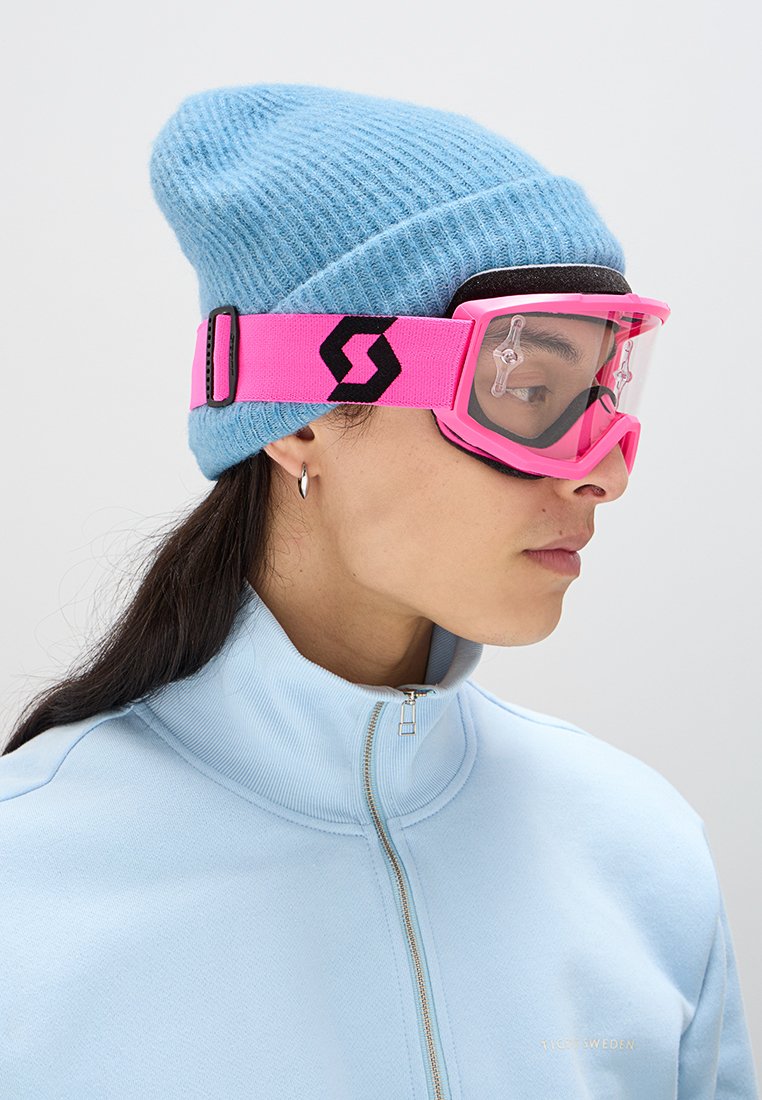 Osoba w niebieskiej dzianinowej czapce, pasującej jasnoniebieskiej kurtce na zamek, srebrnych kolczykach w kształcie kół oraz różowych goglach narciarskich z czarnym paskiem z logo.