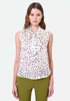 Donna che indossa una camicetta bianca senza maniche con pois scuri e un fiocco annodato al collo, abbinata a pantaloni verde oliva.