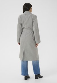 Cappotto lungo grigio in lana con vita regolabile, ampie revers e uno spacco dietro, abbinato a jeans blu a gamba larga e stivaletti neri.