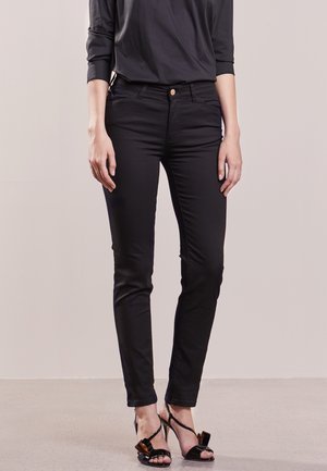 Personne portant un pantalon noir ajusté et un haut noir à manches longues, debout avec les mains détendues le long du corps, sur un sol et un fond gris clair.