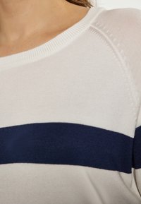 Weißer Pullover mit marineblauen horizontalen Streifen. Weiche, gestrickte Textur und gerippter Kragen. Nahaufnahme zeigt die Detailstiche.