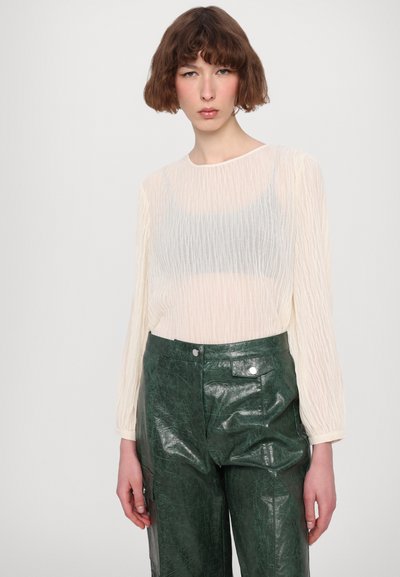 Lichtcrème blouse met lange mouwen en een gekreukte textuur, gecombineerd met donkergroene high-waisted nepleer pants met zakken en een glanzende afwerking.