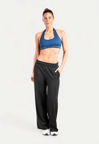 Brassière de sport bleue avec logo, associée à un pantalon de survêtement noir ample avec poches latérales et taille élastique, portée avec des chaussures de sport blanches.