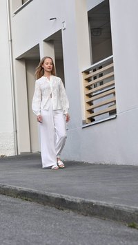 White, patterned blouse with voluminous sleeves, paired with wide-leg white pants and sandals. Background features a textured, gray pavement.
