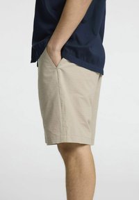 Calções bege com um acabamento texturizado, bolsos laterais e um corte relaxado, usados com uma camisa de manga curta azul escura.
