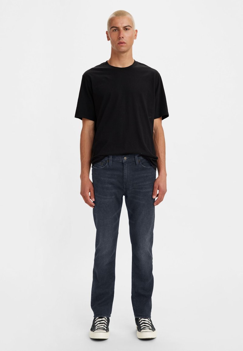 T-shirt noire, jeans slim-fit bleu foncé et baskets montantes noires. Le mannequin se tient droit contre un fond blanc uni.