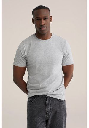 Mand med mørk hud iført en simpel lysegrå t-shirt og mørke jeans, stående med hænderne bag ryggen mod en neutral baggrund.