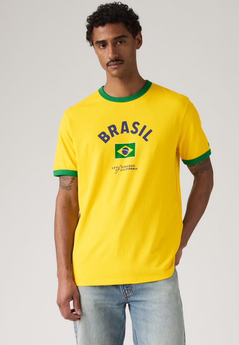 Homme portant un t-shirt jaune avec des bordures vertes, le texte "BRASIL", le drapeau du Brésil et le logo Levi Strauss, accompagné d'un jean bleu clair.
