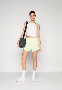 Jauna moteris, vilkinti baltą be rankovių marškinėlį, šviesiai geltonas šortus, baltus sportinius batelius ir kojines, su juodu petine krepšiu, stovinti ant vienodo fono.