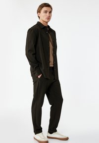 Donkerbruine shirt met een gestructureerd patroon, knoopsluiting aan de voorkant en een zak, gecombineerd met bijpassende broek en witte sneakers met bruine accenten.