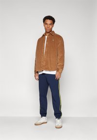 Brown corduroy button-up shirt over a white t-shirt, paired with navy track pants featuring yellow side stripes and white sneakers.