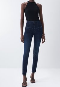 Jeans de tiro alto en azul oscuro con una textura suave de denim, costuras claras y bolsillos frontales. Llevados con un top halter negro con canalé ajustado.