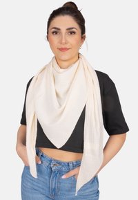 Écharpe beige texturée drapée en forme triangulaire autour du cou, associée à un crop top noir et un jean en denim taille haute.