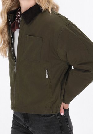 Oliwkowa zielona kurtka z czarnym kołnierzem, przednią kieszenią na piersi i zapinanymi na zamek bocznymi kieszeniami, noszona na białym topie i ciemnych dżinsach.