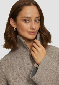 Veste en polaire zippée marron clair avec un col haut, texture douce et motif tissé subtil ; comprend une fermeture éclair métallique et des poignets larges.
