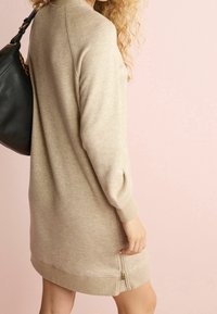 Robe pull en tricot beige avec des manches longues, des poignets côtelés et un détail de fermeture éclair à l'ourlet. Texture lisse sur un fond pastel.
