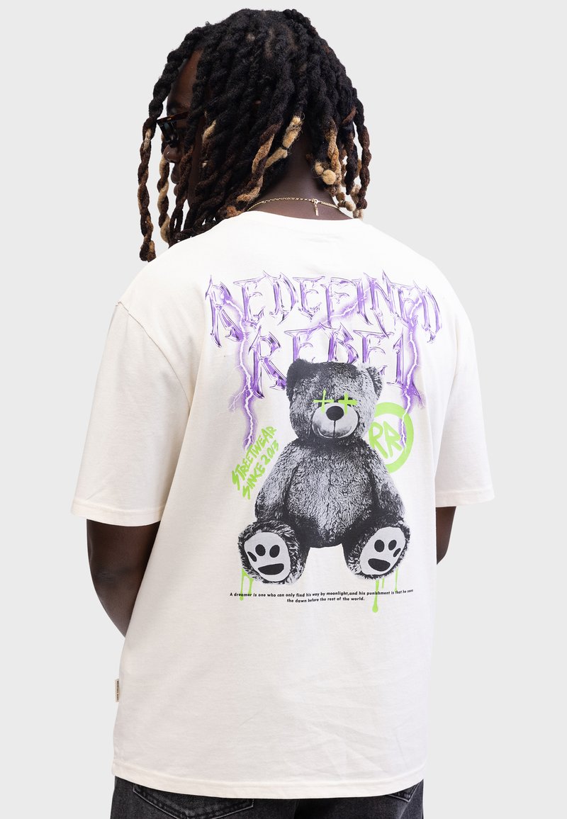 Cremfarbendes T-Shirt mit einem Grafikdruck eines Bären, der grüne Akzente aufweist und lila Text mit der Aufschrift "REDEFINING REBEL" trägt. Kurze Ärmel und lockere Passform.