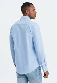 Camisa de manga larga azul claro en una tela suave. Presenta un cuello clásico, puños con botones y un dobladillo curvado con sutiles detalles de costura.