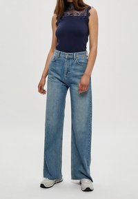 Top sans manches bleu marine avec un bord en dentelle, associé à un jean denim bleu clair à jambes larges. La modèle porte des baskets grises et se tient devant un fond neutre.