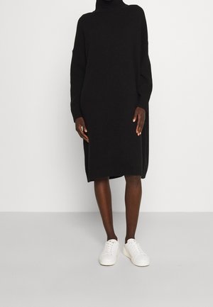 Person trägt ein langes schwarzes Rollkragenpulloverkleid und weiße Sneakers und steht vor einem schlichten hellen Hintergrund.