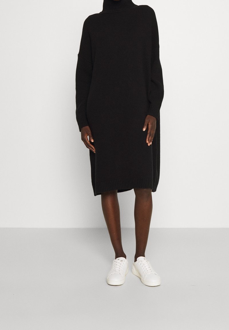 Personne portant une longue robe-pull noire à col roulé et des baskets blanches, debout devant un fond clair uni.