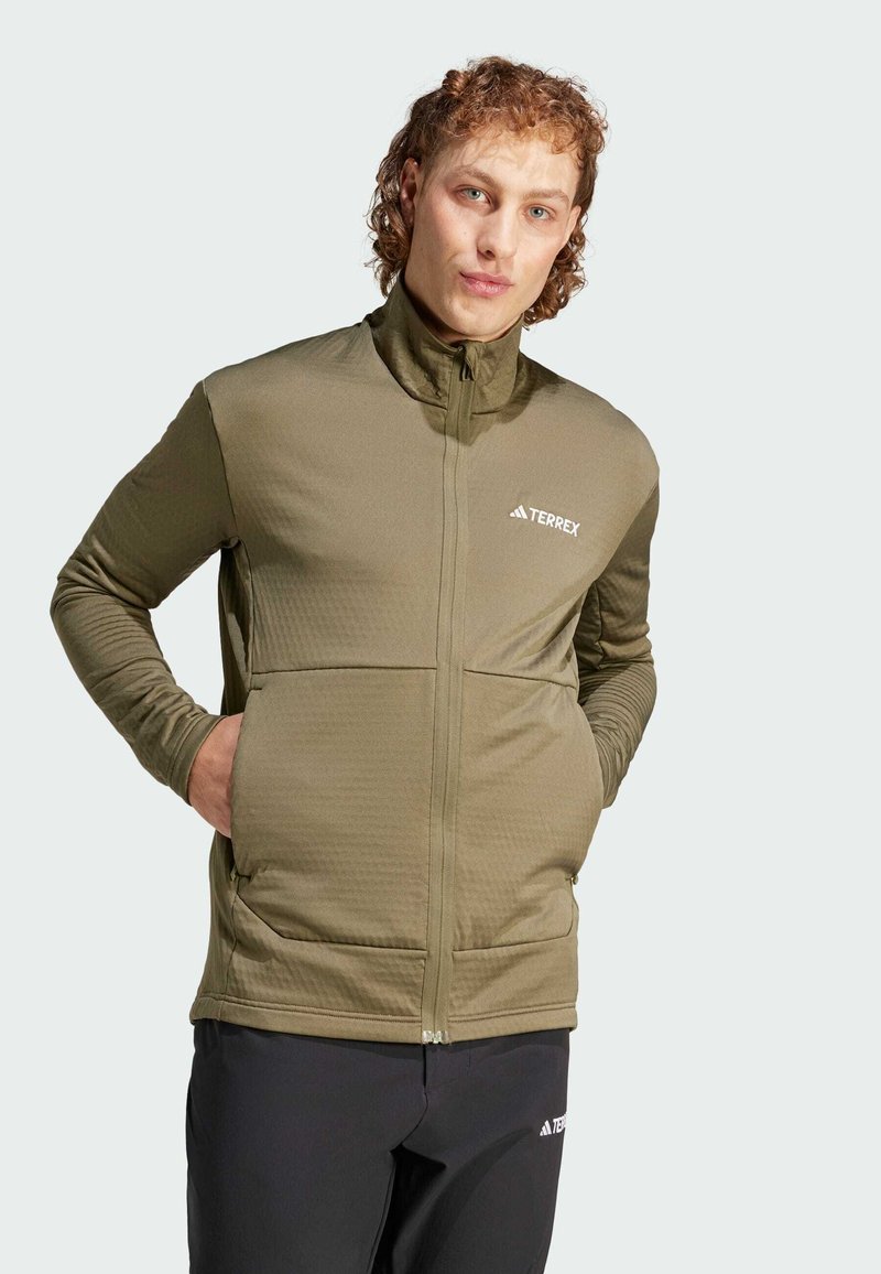 Giacca Terrex verde oliva con colletto alto, chiusura con zip, tessuto texturizzato e tasche frontali. Abbinata a pantaloni neri con branding.