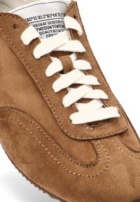 Zapatilla de ante marrón con cordones blancos, puntera redondeada, costuras visibles, superficie texturizada y etiqueta en el interior de la lengüeta.