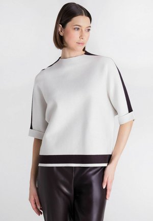Vrouw met kort bruin haar draagt een witte top met zwarte afwerking en een donkere leren broek, poserend met de hand op de heup tegen een witte achtergrond.