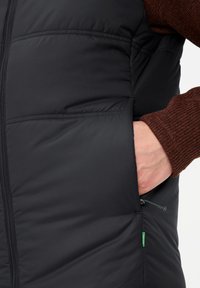 Schwarze gepolsterte Weste mit horizontalen Nähten, Reißverschluss vorne, Seitentasche und grünem Akzent. Strukturierter brauner Pullover darunter sichtbar.