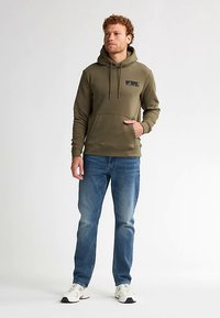 Olivgrüner Hoodie mit einer Fronttasche, Kordelzügen und einem kleinen Logopatch, kombiniert mit blauen Jeans und weißen Sneakers.