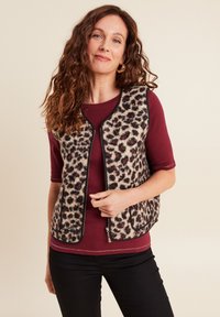 Fleece mouwloze vest met luipaardprint en zwarte afwerking, met rits, en zijzakken, gedragen over een bordeaux top en zwarte broek.
