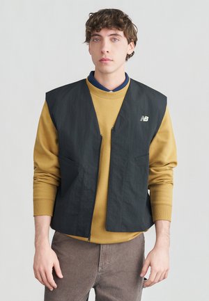 Junger Mann mit lockigem Haar trägt einen senffarbenen Pullover unter einer schwarzen Reißverschlussweste und braune Hose, steht vor einem schlichten hellen Hintergrund.