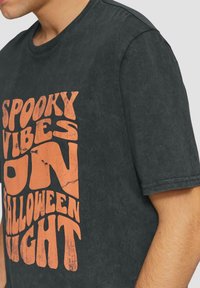 Donkergrijze katoenen t-shirt met vet, versleten oranje tekst: "SPANNENDE SFEER OP HALLOWEEN NACHT." Korte mouwen en een ronde hals.