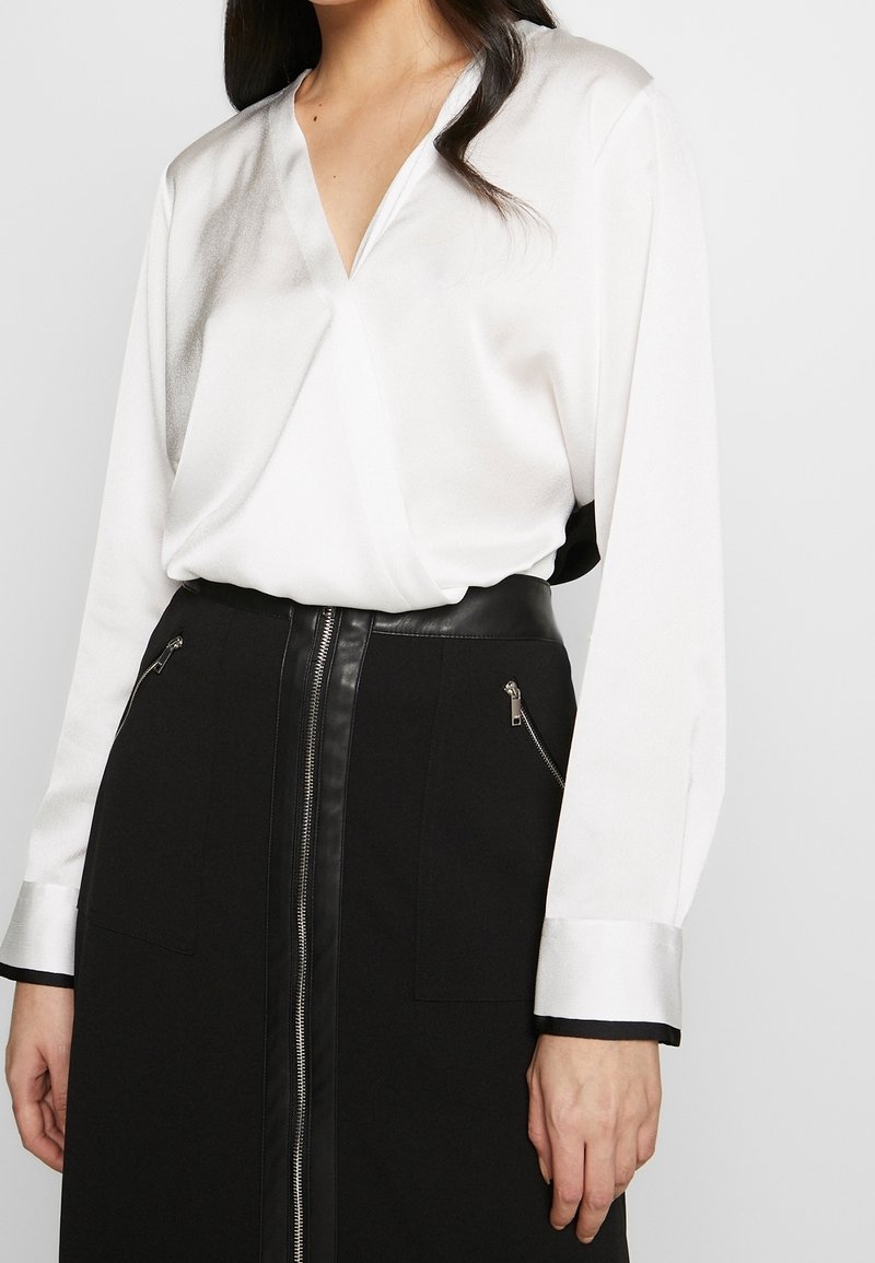 Woman wearing a white long-sleeve V-neck blouse tucked into a high-waisted black skirt with a central zipper and zippered pockets.
