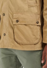 Béžová bavlněná bunda s dvěma předními kapsami, zakulacenými klopami a knoflíkovým zapínáním. Obsahuje diskrétní branding nad kapsou. Hladká textura.