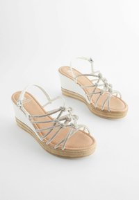 Sandales compensées avec tige en synthétique blanc, brides argent métallisé, semelle intérieure rembourrée marron clair et semelle en caoutchouc texturé aux tons neutres.