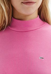 Rosa ribbad polokrage med figursydd design. Litet logotypmärke i röd, vit och svart på bröstet. Släthet och elastiskt tyg.