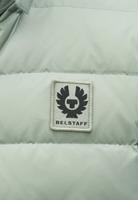 Polstret lysegrøn jakke med et sort broderet mærke med et logo og vingedesign, syet på en glat stofoverflade.