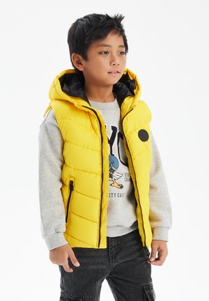 Gul pufferväst med huva, som har svart foder, fickor med dragkedja och en stor svart cirkulär detalj på framsidan. Bärs över en grå sweatshirt.