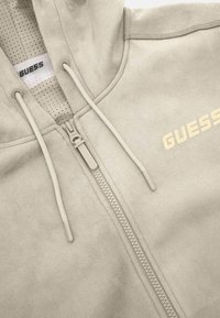 Bežinė zip-up gobtuvė su tekstūruota medžiaga, reguliuojamomis raišteliais ir auksiniu spaudiniu kairėje krūtinės pusėje. Išklota perforuota medžiaga.