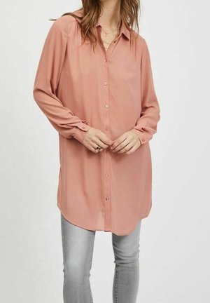 Woman wearing a long-sleeve, buttoned dusty pink blouse paired with light gray skinny jeans, hands clasped in front.