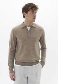 Suéter beige con cierre, cuello tipo sacerdote, textura de punto, mangas largas y puños acanalados, usado sobre una camisa blanca, combinado con pantalones claros.