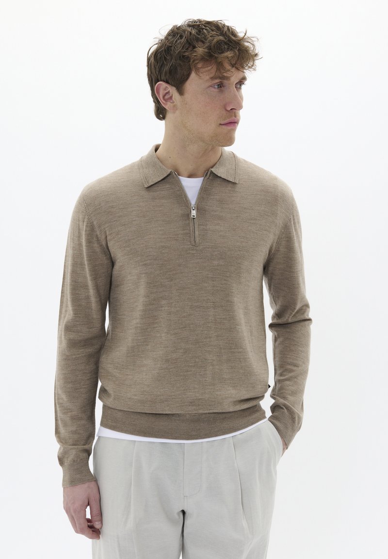 Suéter beige con cierre, cuello tipo sacerdote, textura de punto, mangas largas y puños acanalados, usado sobre una camisa blanca, combinado con pantalones claros.