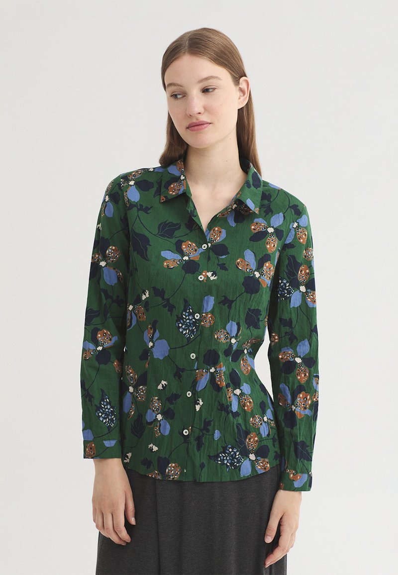 Femme portant un chemisier vert à manches longues boutonné avec des motifs floraux bleus et bruns, debout devant un fond clair uni.